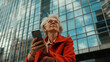 © yanapopovaiv - an elderly woman with gray hair, holding a phone against the background of high-rise windows, sitting near a skyscraper with glasses, a retired businesswoman
