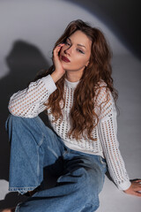 Wall Mural - Fashion beautiful woman in a stylish white sweater with jeans sits on the floor in the studio