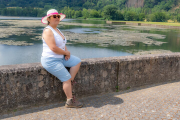 Wall Mural - Senior female tourist sitting on fence at shore of Echternach lake, serene expression, hat, shorts and sunglasses, calm water and green trees in background, sunny summer day in Luxembourg