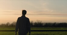 Young Man Walking In A Field Towards The Sun. Silhouette Man Walks Along The Grass Field At Sunset. A Boy Walking On Sunset Background. Rural Landscape View. Agricultural Concept.