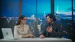 © kinomaster - Male and female presenters in the studio. Man and woman news hosts sit at desk presenting evening events, talking joking on air, virtual graphic background at back.