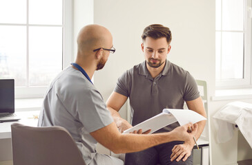 Portrait of a young man patient in medical office listening to a friendly doctor holding report file with appointment and giving consultation during medical examination in the clinic.