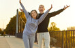© Studio Romantic - Senior couple having fun during outdoor fitness workout on summer morning. Happy old man and woman in sportswear standing on bridge, feeling full of energy, raising hands up and smiling. Sport concept
