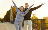 Fototapeta Natura - Senior couple having fun during outdoor fitness workout on summer morning. Happy old man and woman in sportswear standing on bridge, feeling full of energy, raising hands up and smiling. Sport concept