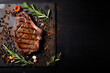 Grilled beef steak with spices, herbs, seasoning on dark black wooden board background, top down view, text copy spacer.