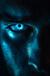 Glowing blue eye of a mysterious man. Serious expression. Evil man in the dark. Intense gaze. Paranormal glowing eye concept. Closeup of glowing evil eyes in the dark. Halloween creepy concept. 