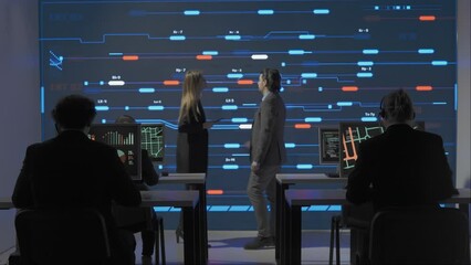 Technical support team in control room. Man and woman specialists discussing work, digital screen shows infrastructure monitoring infographic network data.