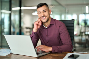 Happy young professional Latin business man looking at camera sitting at work desk using computer. Portrait of smiling male company worker employee, project manager or entrepreneur in office.