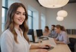 © kotlyarn - A young woman in a casual shirt works at her desk, colleagues in the background. She gazes confidently towards the camera.