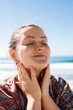© Karina Allakhverdian/Stocksy - A young woman touching her face and smiling at a beach