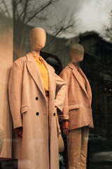 Wall Mural - Trendy stylish mannequins in a store window dressed in seasonal fashion cloth. Beige long coat and a bright yellow shirt.
