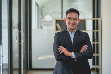 Wall Mural - Asian businessman in a suit is smiling and standing in a room with a white background. He is wearing glasses and has his arms crossed