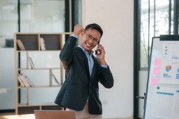 Wall Mural - Asian businessman in a suit is talking on his cell phone while standing in front of a white board with graphs on it