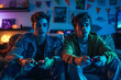 Two young men sitting on a sofa holding game controllers and playing videogames, blue lighting in the room