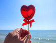 A stick in the shape of a volumetric red heart in woman hand against background of blue sea, sandy beach and blue sky on sunny summer day. Concept love, Valentine Day, falling in love. Vertical
