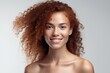 © Iona - Beautiful young woman with curly red hair and freckles, smiling naturally on a light background.
