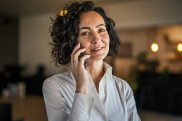 Wall Mural - One woman mature caucasian female businesswoman entrepreneur stand at work or home use mobile phone making a call talk real people copy space wear white shirt curly hair happy smile