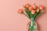 Fototapeta Tulipany - Vibrant orange tulips arranged in a clear glass vase held by hands, set against a soft pink background. Concept of spring, freshness, and floral decor