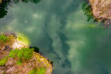 Fototapeta  - Położone wśród lasów jezioro, którego woda ma szmaragdowy kolor. Brzegi pokrywają żółte, suche trawy, bezlistne drzewa, między którymi widać zielone korony drzew iglastych. 