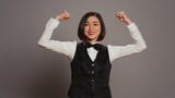 Fototapeta Tulipany - Asian hotel concierge flexing arms muscles in grey studio, showing off her strength and power in front of camera. Receptionist with bow and uniform standing with biceps flexed. Camera B.