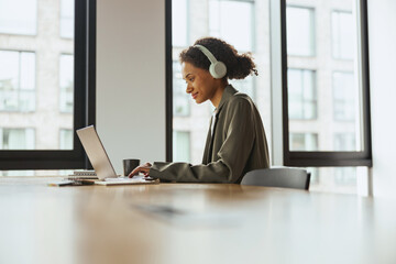 A woman with headphones sits at a table using a laptop computer in nice modern coworking office