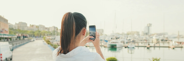 Wall Mural - Young woman taking photo of the sea port on mobile phone. Back view, Panorama