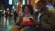 An African American man and woman, smiling and laughing, sit on a bench on a chilly night, wrapped in thick winter coats, engrossed in their phones under the glow of warm streetlights.