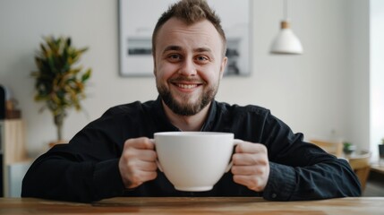 Wall Mural - smiling man with a big smile in a modern light interior sits at the table and holding a very large white cup of coffee 