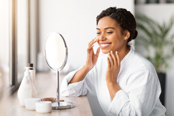 Sticker - Happy young black woman applying facial moisturizer while looking in makeup mirror