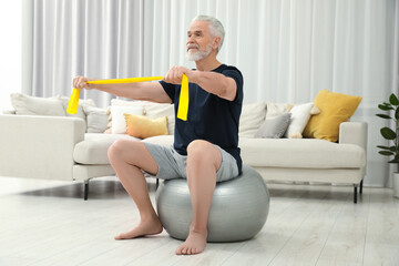 Wall Mural - Senior man doing exercise with elastic resistance band on fitness ball at home