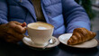 Woman holding a cup of cappuccino and a croissant in a plate nearby on the table