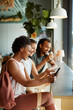© Flamingo Images - Three diverse friends sit at a high table by a window in a cafe after a yoga lesson. They drink coffee and look at mobile while laughing together