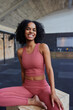 © Flamingo Images - A young black fit woman sitting on box in a gym and smiles at the camera in pink sportswear