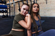 © Flamingo Images - Two confident diverse women sit together on a gym floor in their workout clothes after a fitness class