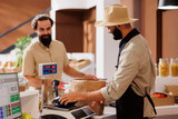 Fototapeta Tulipany - Middle eastern salesman being of service to caucasian customer at checkout counter. At cashier desk, male vendor wearing a hat while using digital scale to measure items for the young client.