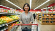 Beautiful middle-aged Japanese woman smiles while shopping at the supermarket with her cart in the fruit section.