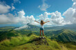 Female traveler with arms outstretched standing on a mountain summit overlooking green hills and clouds. Young woman cheering arms up of joy