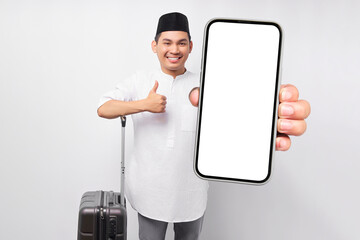 Wall Mural - cheerful young Asian Muslim man standing while holding a suitcase and smartphone, showing thumb up gesture isolated on white background. Ramadan and eid Mubarak concept