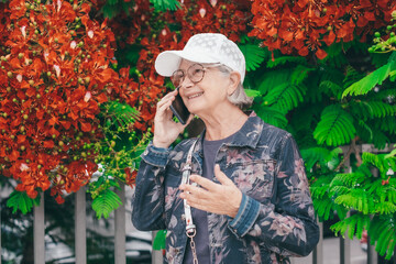 Wall Mural - Attractive senior woman in jacket jeans and white cap walking in spring park outdoors using mobile phone talking and gesturing. People urban lifestyle concept