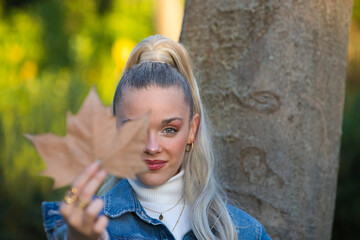 Pretty young blonde woman with a ponytail in her hair covers one eye with a dry leaf. Focus on the face. Autumn and winter concept.