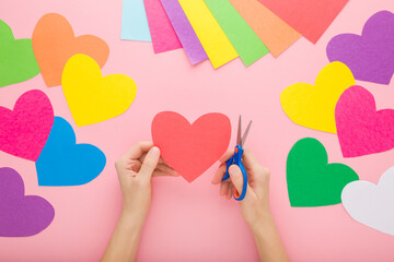 Wall Mural - Young adult woman hands holding scissors and cutting red heart shape from application paper on pastel pink table background. Making decoration elements. Closeup. Point of view shot. Top down view.