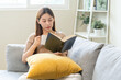 © KMPZZZ - Having relaxing weekend, comfort asian young woman with cup of beverage, drinking coffee or tea of mug. Girl sitting on sofa open reading book enjoy of rest after get up in morning lifestyle at home.