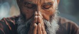 Fototapeta  - A man with facial hair and a beard is making a praying gesture with his hands folded in front of his face. The wrinkles on his forehead show his concentration