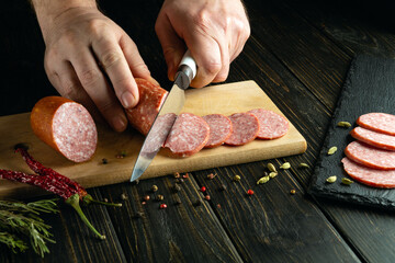 Wall Mural - Chef hands with a knife cut sausage on the kitchen table for preparing meat snacks in a tavern.