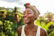 A beautiful young black woman with a colorful headband smiles and laughs with her eyes closed. She has a white T-shirt. In the background is a tropical garden with palm trees