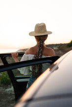 Woman traveler, wearing a straw hat, enjoys the sunset from her car during a road trip