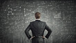 Businessman rear view with hands on hips thinking in front of a blackboard background written with different formulas and equations