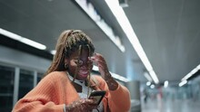 Joyful Young African Woman With Vitiligo Laughing While Looking At Smartphone In Subway Station. Dressed In Orange Cardigan Female Feel Moment Of Genuine Happiness, Light-heartedness In Everyday Life

