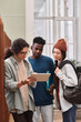 © Mediaphotos - Vertical portrait of diverse group of three young students looking at tablet screen while standing in college library with backpacks