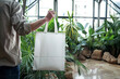 A man holding a blank white tote, reusable shopping bag in a dynamic, modern atrium filled with plants, underlining the integration of nature into architectural design,